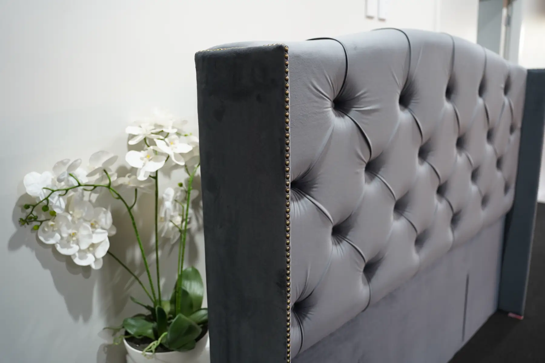 A grey velvet headboard, deep buttoned with a studded border, curving into a tall wing.